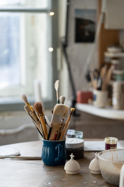 Diversity of paintbrushes put into blue mug standing on round wooden table with pottery equipment