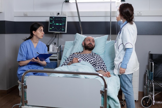 Doctor adjusting intravenous medication iv drip line flow for middle aged man recovering after surgical intervention. Nurse sitting at patient bedside writing medical observations on clipboard.