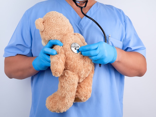 Doctor in blue uniform and white latex gloves holding a brown teddy bear