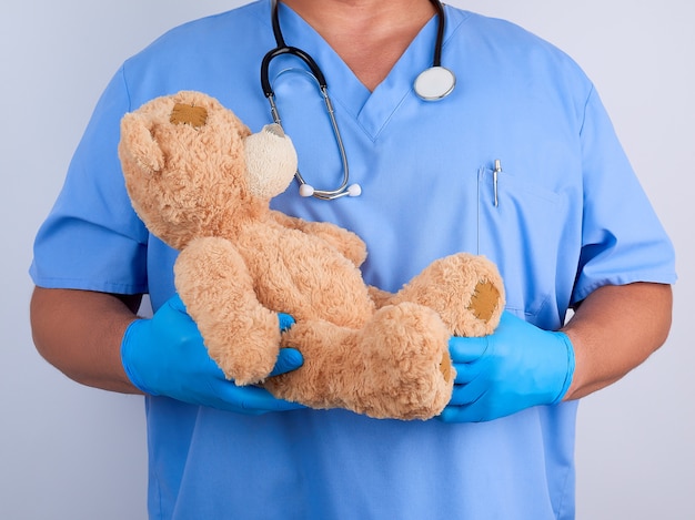 Doctor in blue uniform and white latex gloves holding a brown teddy bear