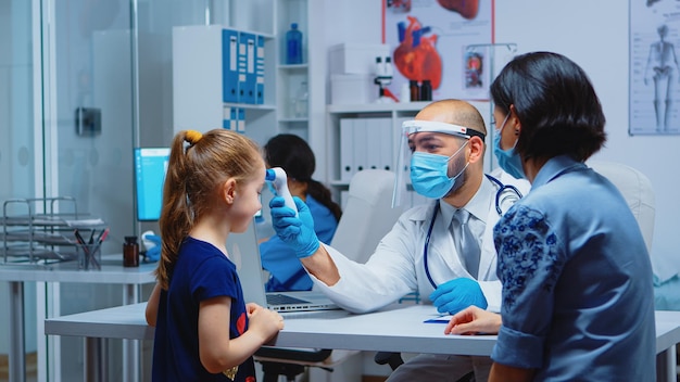 Doctor checking child temperature wearing protection mask during covid-19. Pediatrician specialist in medicine with visor providing health care services, consultation, treatment in hospital cabinet.