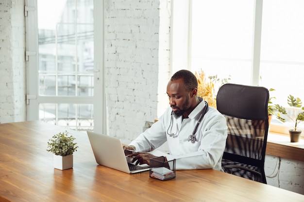 Doctor consulting for patient, working with laptop. African-american doctor during his work with patients, explaining recipes for drug. Daily hard work for health and lives saving during epidemic.