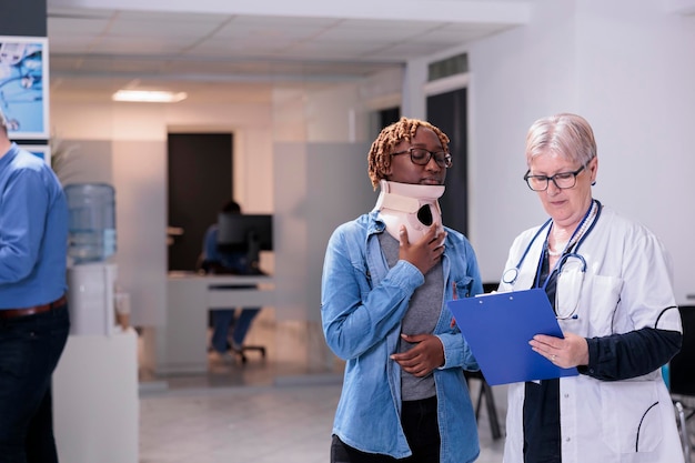 Doctor examining woman with neck collar, wearing cervical foam brace after accident injury. Diverse people doing checkup visit consultation with medical appointment in waiting area lobby.