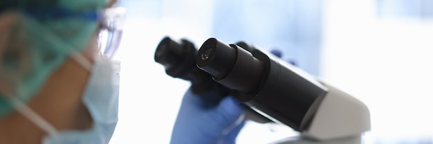 Doctor in glasses and a medical mask looks through microscope.