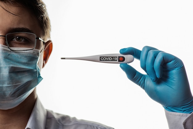 A doctor in a glasses and surgical mask holds an electronic thermometer to measure temperature. Signs of flu COVID 19 Coronavirus