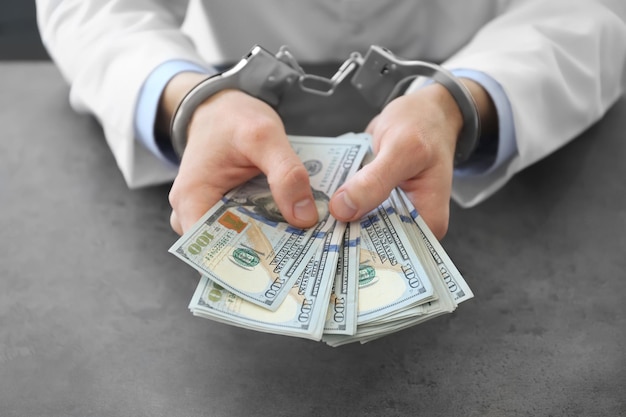 Doctor in handcuffs holding bribe at workplace closeup