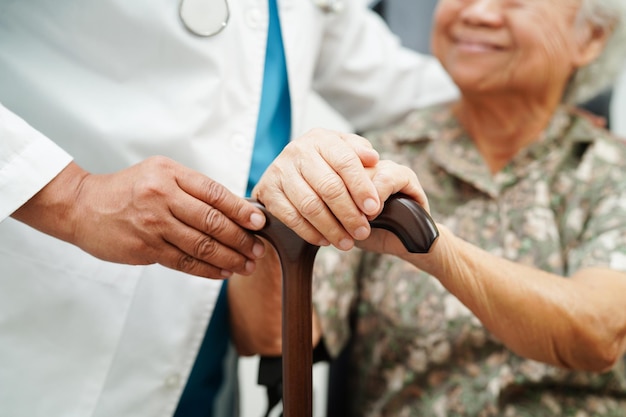 Doctor help Asian elderly disability woman patient holding walking stick in wrinkled hand at hospital