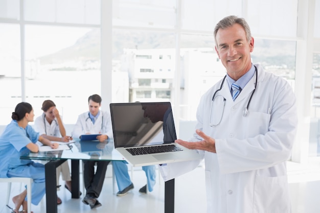 Doctor holding laptop with group around table in hospital