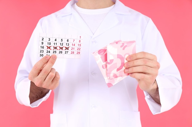 Doctor holds menstrual calendar and pads on pink background