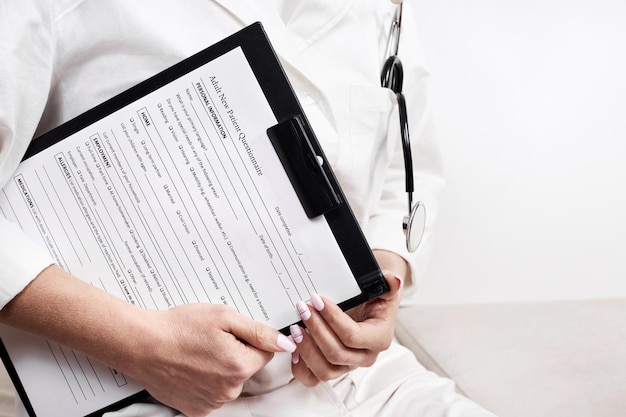 A doctor in a medical gown and with a phonendoscope holds a planchette in his hand with an Adult New Patient Questionnaire