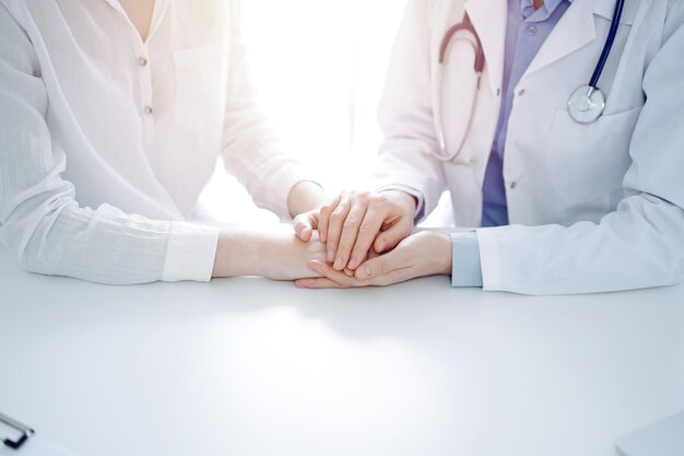 Doctor and patient sitting near each other at the table in clinic office. The focus is on female physician's hands reassuring woman, only hands, close up. Medicine concept