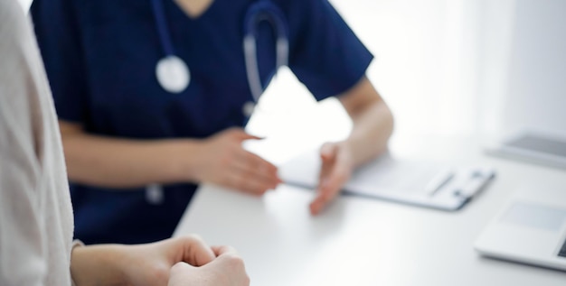 Doctor and patient sitting at the table in clinic while discussing something. The focus is on female patient's hands, close up. Medicine concept.