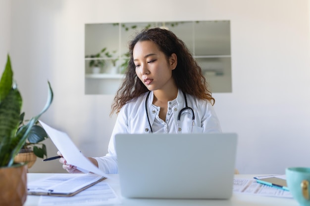 Doctor professional female Asian doctor wearing uniform taking notes in medical journal filling documents patient illness history looking at laptop screen student watching webinar