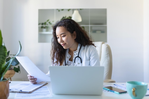 Doctor professional female Asian doctor wearing uniform taking notes in medical journal filling documents patient illness history looking at laptop screen student watching webinar