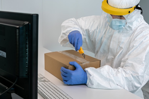 Doctor on protective suit and plastic face shield opens a box of face masks during Covid 19. Contaminated mail