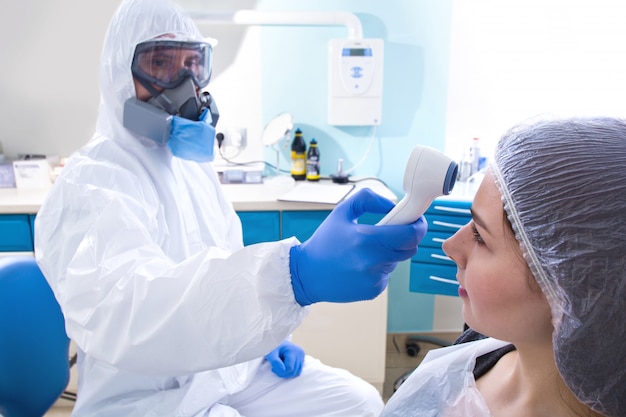 Doctor in protective suit uniform and mask checking temperature of young female patient.