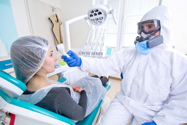 Doctor in protective suit uniform and mask checking temperature of young female patient.