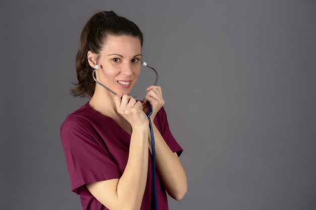 Doctor in purple scrubs putting on stethoscopes looking at camera on gray background