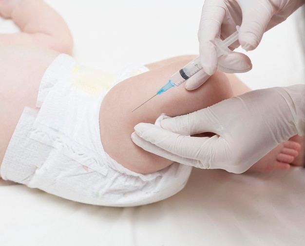 The doctor's hands in rubber protective gloves hold a syringe. An infant receiving the vaccine in the leg. Visit to the pediatrician in the hospital. A two-month-old baby. To close.