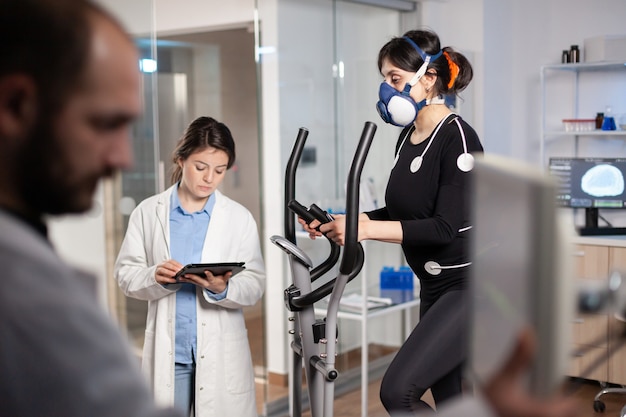 Doctor scientist holding tablet pc and controling EKG data showing on laboratory monitors. Lab science researcher measuring endurance of athlete, heart rate using electrodes and mask.