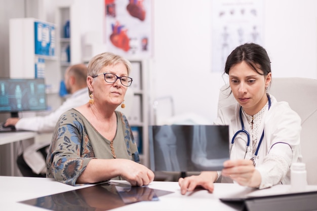 Doctor talking about knee pain with senior patient holding radiography in hospital office . Medic wearing white coat working on computer.