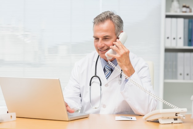 Doctor using laptop and talking on phone at desk