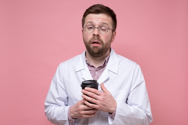 Doctor very tired and looks sleepy he holds paper cup of coffee in hands and tries to open his eyes