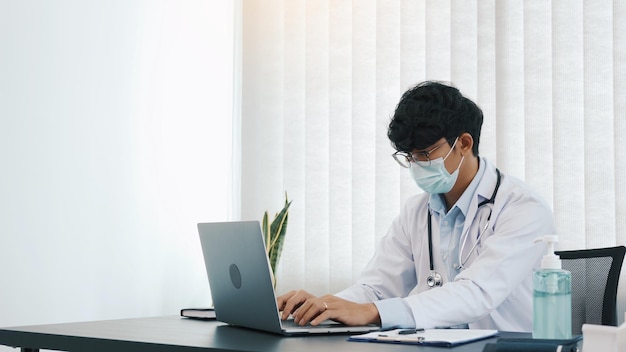 Doctor was sitting in the office working on a laptop computer while wearing a mask