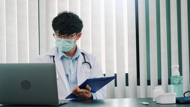 Doctor was sitting in the office working on a laptop computer while wearing a mask