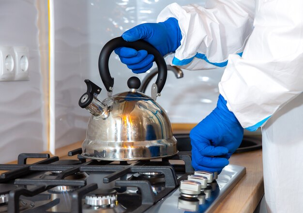 Doctor wearing medical protective suit, goggle, mask, and gloves preparing tea on the kitchen. Teapot on the gas stove. Protection mers by virus epidemic. Coronavirus (COVID-19). Healthcare concept.