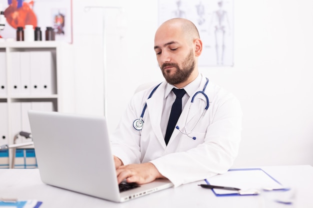 Doctor wearing stethoscope and white coat in hospital office using laptop to write patient treatment. Medical practitioner using notebook in clinic workplace , confident, expertise, medicine.