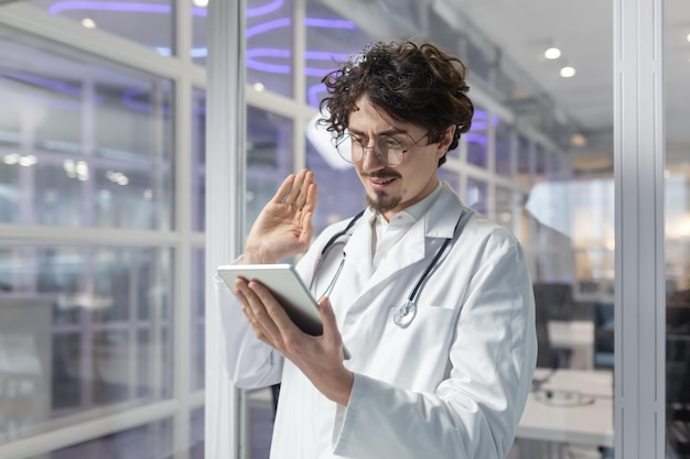 A doctor wearing a white lab coat and stethoscope holds a tablet inside a medical office of a clinic