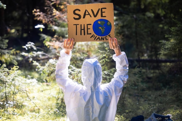 Doctor in a white anti-epidemic suit holding a cardboard banner in hands. In the woods.