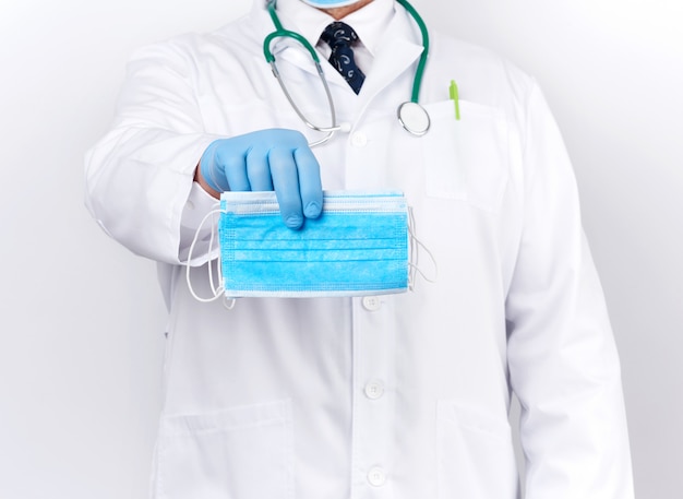 Doctor in a white coat, blue latex sterile gloves holds textile medical masks in his hand