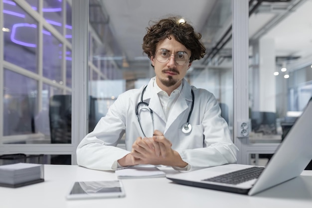 A doctor in a white coat and stethoscope sitting at a desk inside a medical office using a laptop