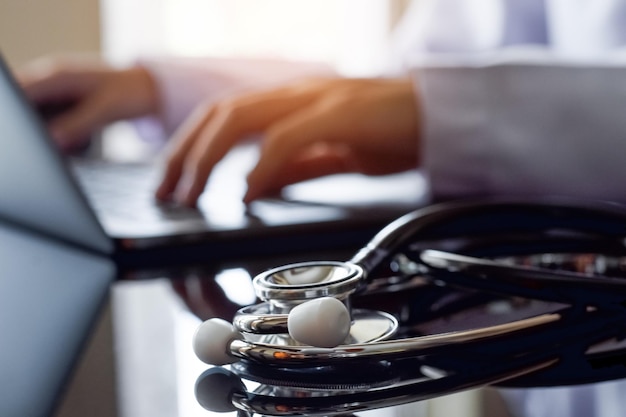 Doctor in white lab coat typing on laptop computer with medical stethoscope on the desk.