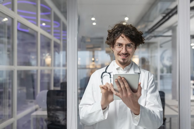 A doctor in a white medical coat and stethoscope holds a tablet for a medical consult in his office