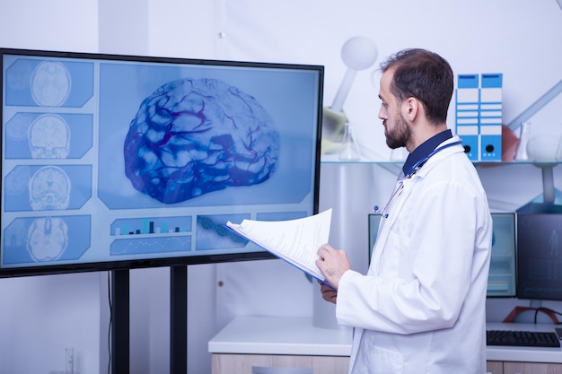 Doctor with clipboard on his hand looking at a brain on a big monitor. Confident doctor. Doctor in uniform.