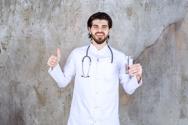 Doctor with a stethoscope holding a glass of pure water and showing positive hand sign. 