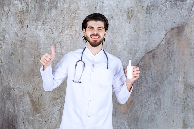 Doctor with a stethoscope holding a white tube of hand sanitizer spray and showing thumb up sign