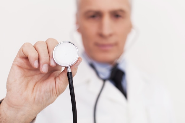 Doctor with stethoscope. Senior doctor in uniform looking at camera and holding his stethoscope outstretched while standing isolated on white