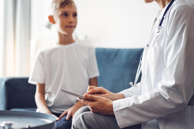 Doctor woman and kid boy patient at home. The pediatrician using tablet computer while filling up medical records, close up. Medicine, healthcare concepts