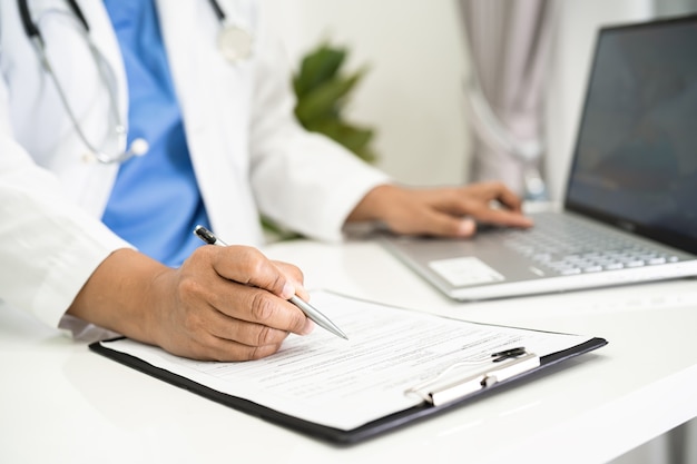 Doctor write health medical note in clipboard with laptop in hospital.