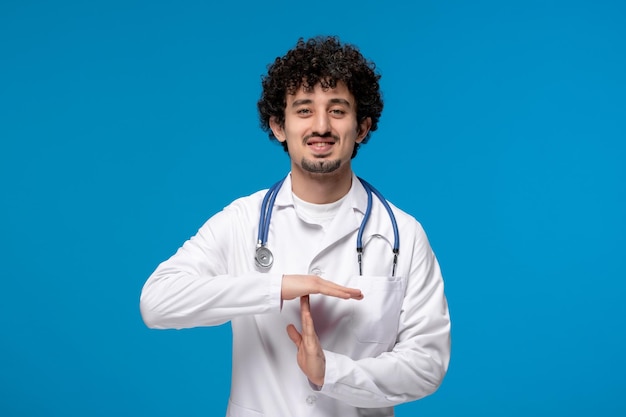 Doctors day curly brunette cute guy in medical uniform showing stop sign and smiling