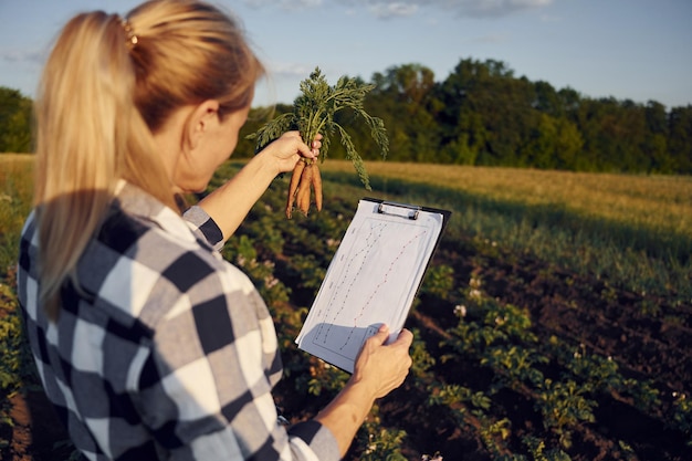 Document with information and carrots in hand Woman is on the agricultural field at daytime