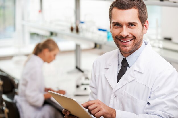 Documenting the result of experiments. Smiling young male scientist holding digital tablet and looking at camera while his female colleague working in the background