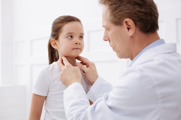 Does it hurt. Determined gentle prominent doctor examining little girls glands while comparing her symptoms to diagnosis