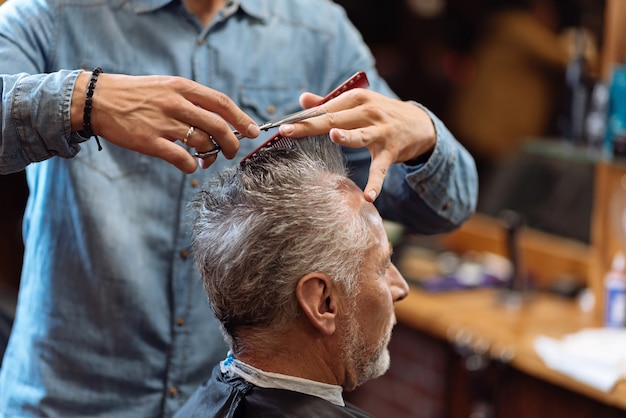 Doing accurately. Close-up of male hairdresser holding scissors and comb while doing haircut to senior man.