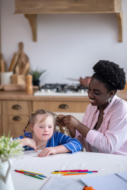 Doing hair. A smiling dark-skinned woman doing hair to the cute girl with down syndrome