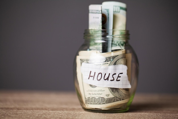 Dollar bills in glass jar and text house on wooden table